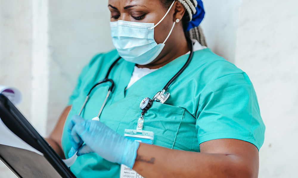 Black woman in mask, scrubs, gloves holding a tablet